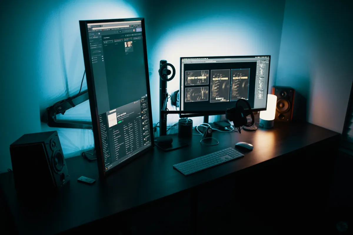 Placa de vídeo integrada vs dedicada: qual a diferença? 2 Dois monitores de computador em ambiente de trabalho. Um deles está colocado na vertical, como se fosse um grande tablet. A mesa é preta, assim como os monitores, e a parede atrás é azul. O ambiente está pouco iluminado. Há outros periféricos, como um teclado e uma caixa de som.