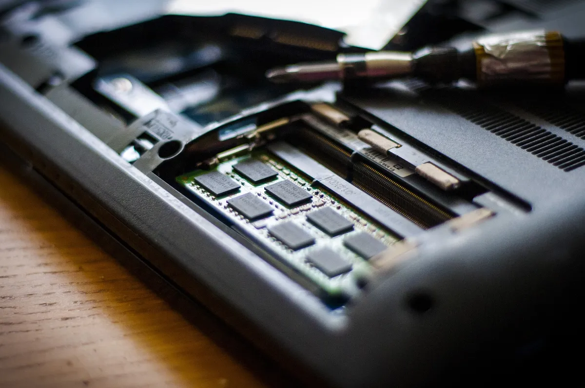 Tipos de Memória RAM: DDR2, DDR3, DDR4, DDR5 - O Guia Completo 4 Comportimento de memórias RAM aberto na parte traseira de um notebook que está passando por manutenção. O notebook é preto, e é possível ver as memórias encaixadas na placa mãe na parte interna. Há uma chave philips pequena, com um remendo de fita adesiva no cabo, próxima ao compartimento aberto.