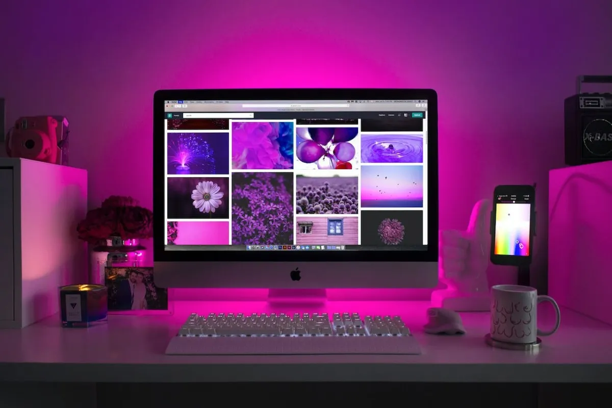 Organização de Cabos e Fios: 7 dicas para deixar seu setup mais clean 2 Monitor e teclado brancos, sobre mesa de trabalho branca, bem organizada. O monitor exibe várias fotos de papel de parede de flores e objetos em tons rosa e lilás. Tudo está iluminado por uma luz rosa intensa.
