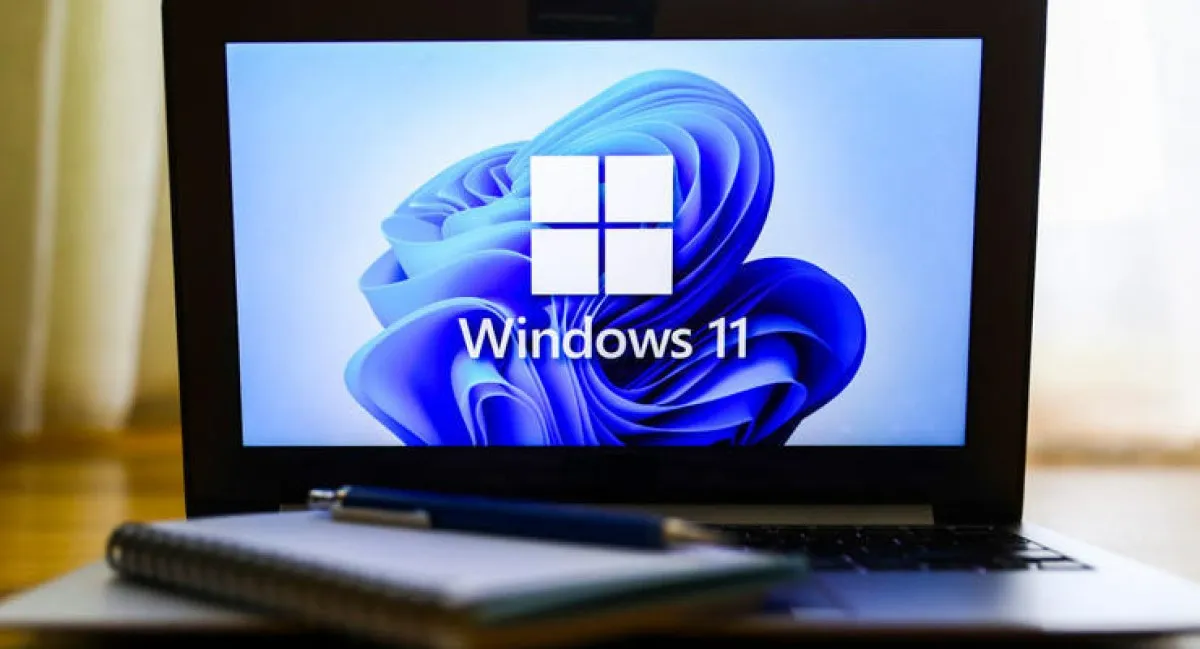 Notebook aberto com o tradicional símbolo de 4 quadrados do Windows sobre o papel de parede rosa azul do Windows 11. Há uma caderneta e uma caneta sobre o teclado.