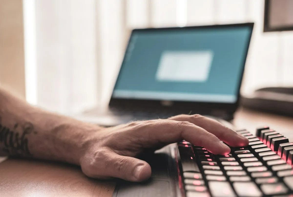 Como limpar computador e deixar seu PC mais rápido (atualizado 2024) 2 Detalhe mostrando uma pessoa digitando com uma mão em um teclado com iluminação vermelha. Não podemos ver o monitor, há um notebook aberto na mesa ao lado.