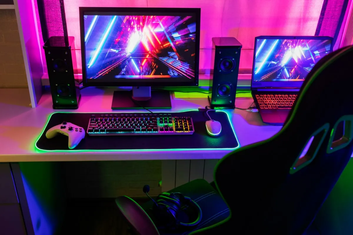 Vista frontal de uma mesa gamer branca, com um monitor gamer e um teclado com luzes multicoloridas, e um notebook gamer do lado, com teclado iluminado em vermelho. O monitor e o notebook tem o mesmo papel de parede multicolorido, com o que parece ser um detalhe do interior de uma nave especial. O espaço é iluminado em uma luz rosa e há uma cadeira gamer de costas, da cor preta.