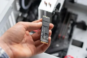 Melhor Pasta Térmica: TOP 10 Melhores (Cooler, Master, Grizzly e mais) 42 Detalhe mostrando a mão de uma pessoa segurando uma unidade SSD M.2. Ao fundo vemos o que parece ser um gabinete aberto. O fundo está desfocado. Imagem representando o melhor SSD para PC gamer.