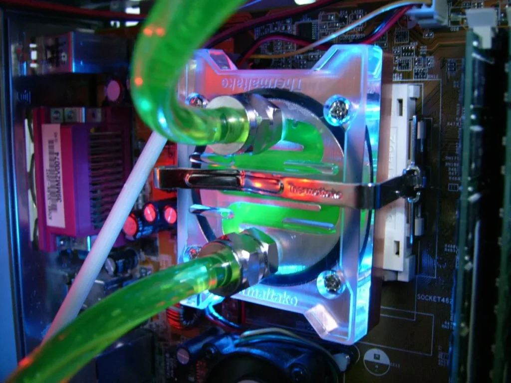 Página Inicial - Guru Recomenda 78 Imagem mostrando um water cooler acoplado acima do processador em gabinete de PC. Há duas mangueiras transparentes saindo do cooler, iluminadas com luz RGB verde claro. A bomba do water cooler também é transparente e iluminada com a mesma cor. Foto representativa do melhor water cooler.