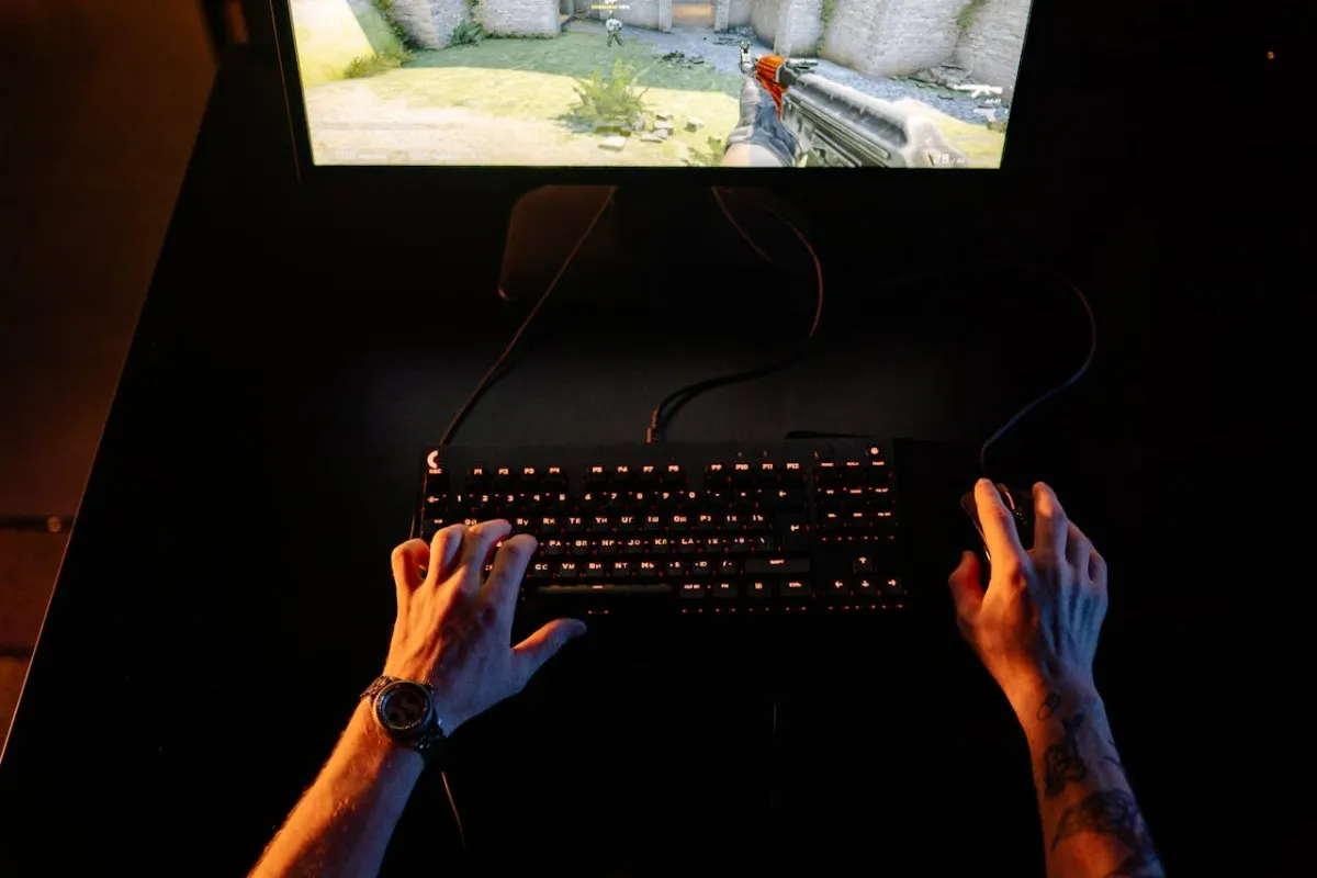 Imagem mostrando duas mãos masculinas de um gamer sobre teclado gamer com luzes RGB vermelhas. É possível uma pequena parte da tela, que apresenta uma paisagem gramada. A mesa é preta e o ambiente está escuro.